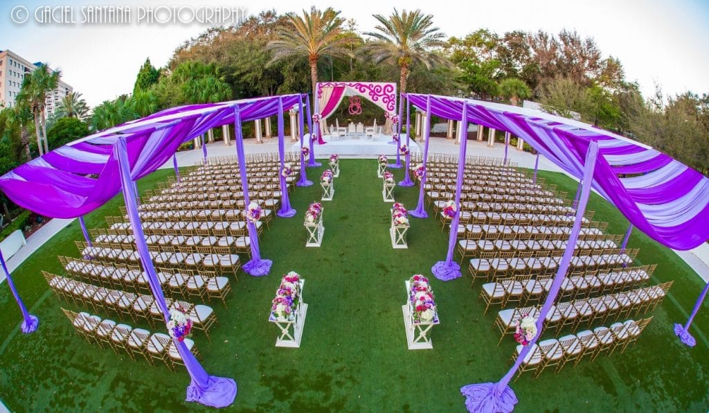 outdoor wedding reception set up on green lawn with palm trees, gold and white chairs, and purple canopies and flowers