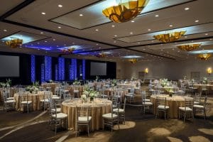 hotel ballroom set up for wedding reception with gold linens, white floral arrangements, and purple lighting