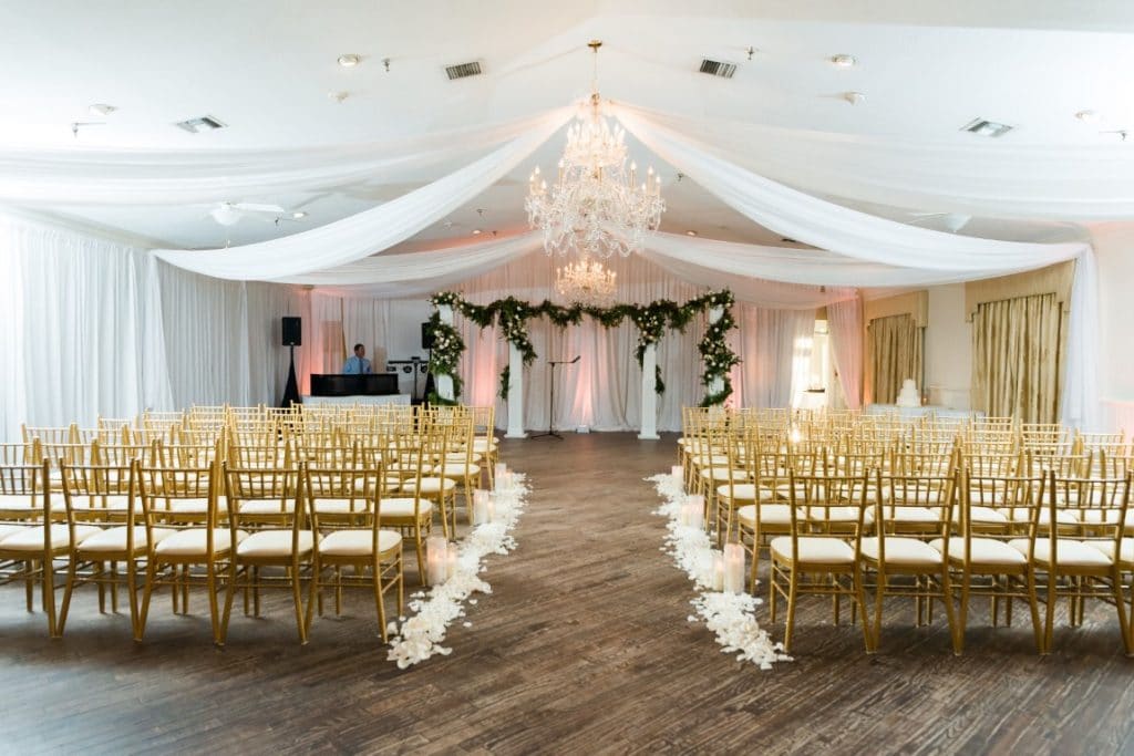 The-Highland-Manor-Ceremony hall decorated with floral arch, gold chairs, white flowers and large chandelier lighting the room.