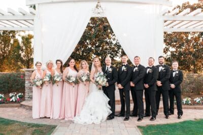 Wedding party portrait at The Royal Crest Room in St. Cloud Florida featuring bridesmaids in blush pink gowns and groomsmen in black tuxedos posing under a draped pergola in the garden venue setting