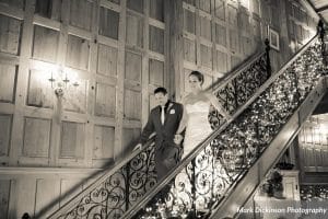 Bride and groom descend an ornate wrought iron staircase with warm candlelit wood-paneled walls at Estate on the Halifax, a historic wedding venue in Port Orange near New Smyrna Beach, Florida, photographed by Mark Dickinson Photography