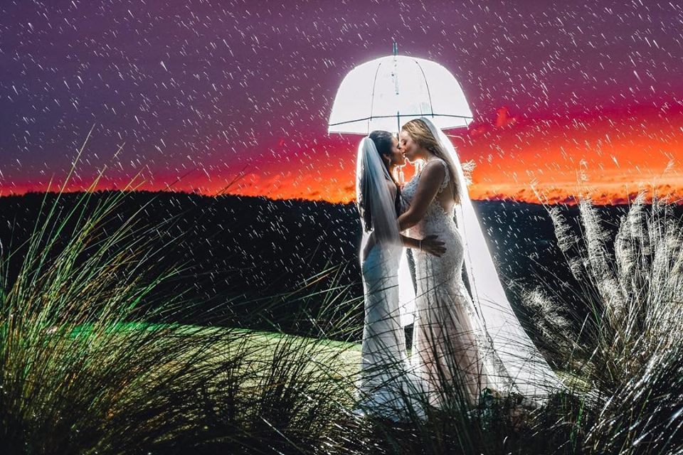 That First Moment - two brides kissing under umbrella in the rain