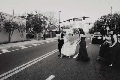 Wedding party crossing a downtown Orlando street intersection in black and white, bride and groom leading bridesmaids in dark gowns near 1010 West historic venue in Orlando Florida