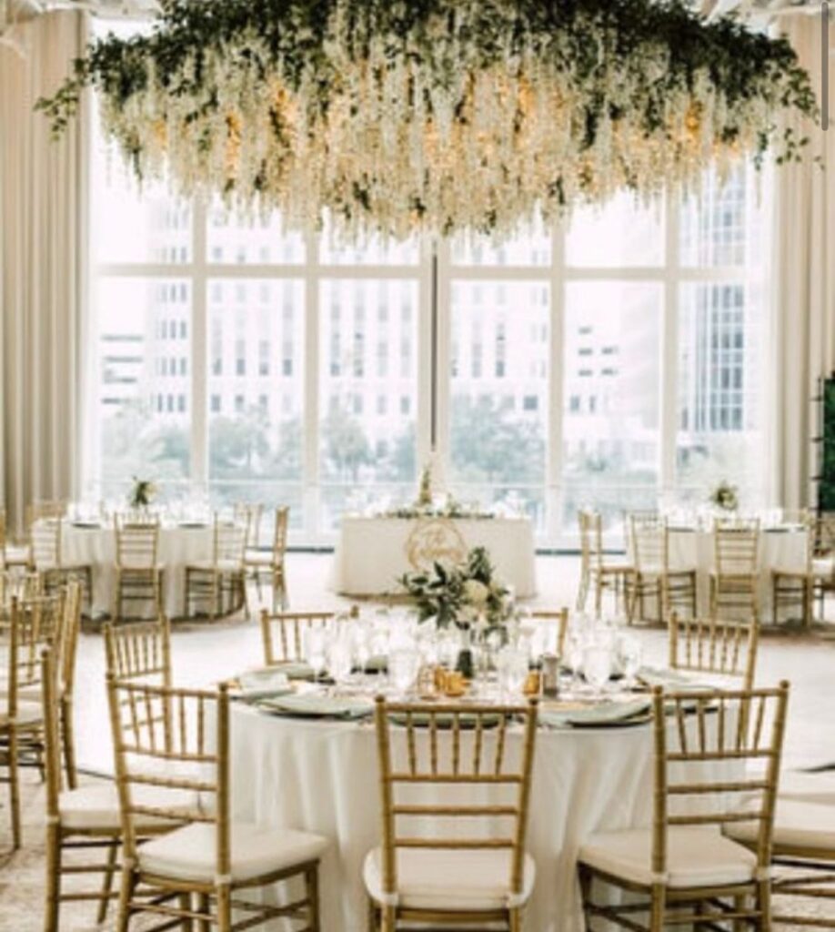 room set up for wedding reception with round white tables, gold chairs, and large white floral chandelier hanging from ceiling