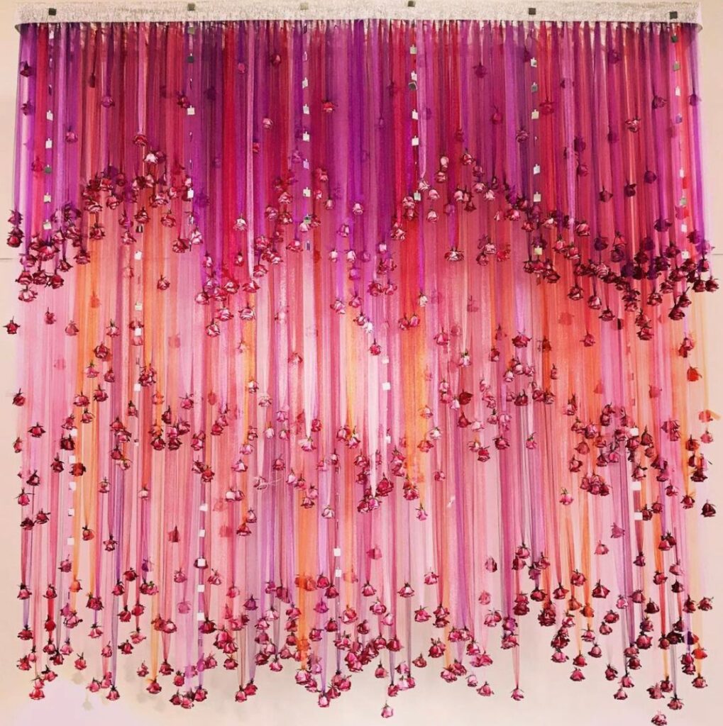 wall decor made from a curtain of pink and orange ribbons with flowers hanging at the ends