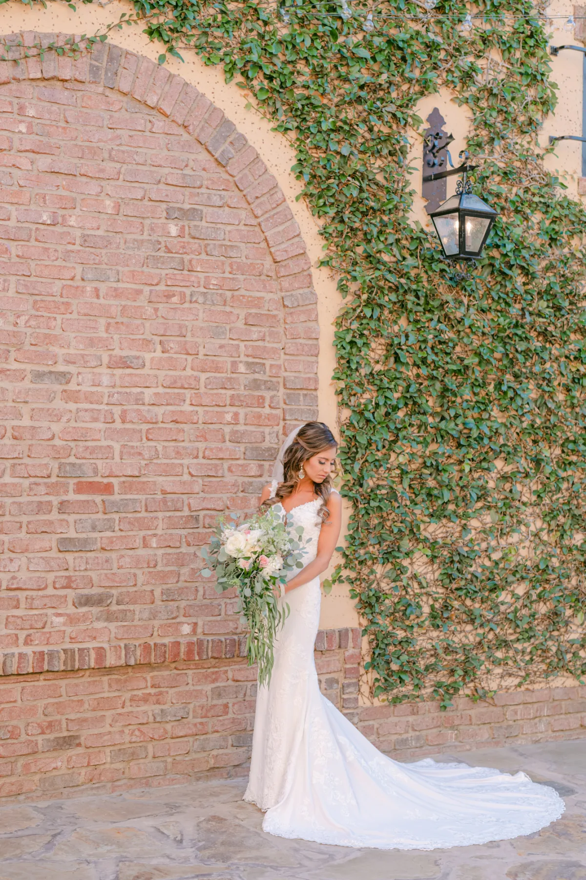 Bride in a lace wedding dress holding a bouquet, standing by an ivy-covered wall and brick arch at Bella Collina in Florida.