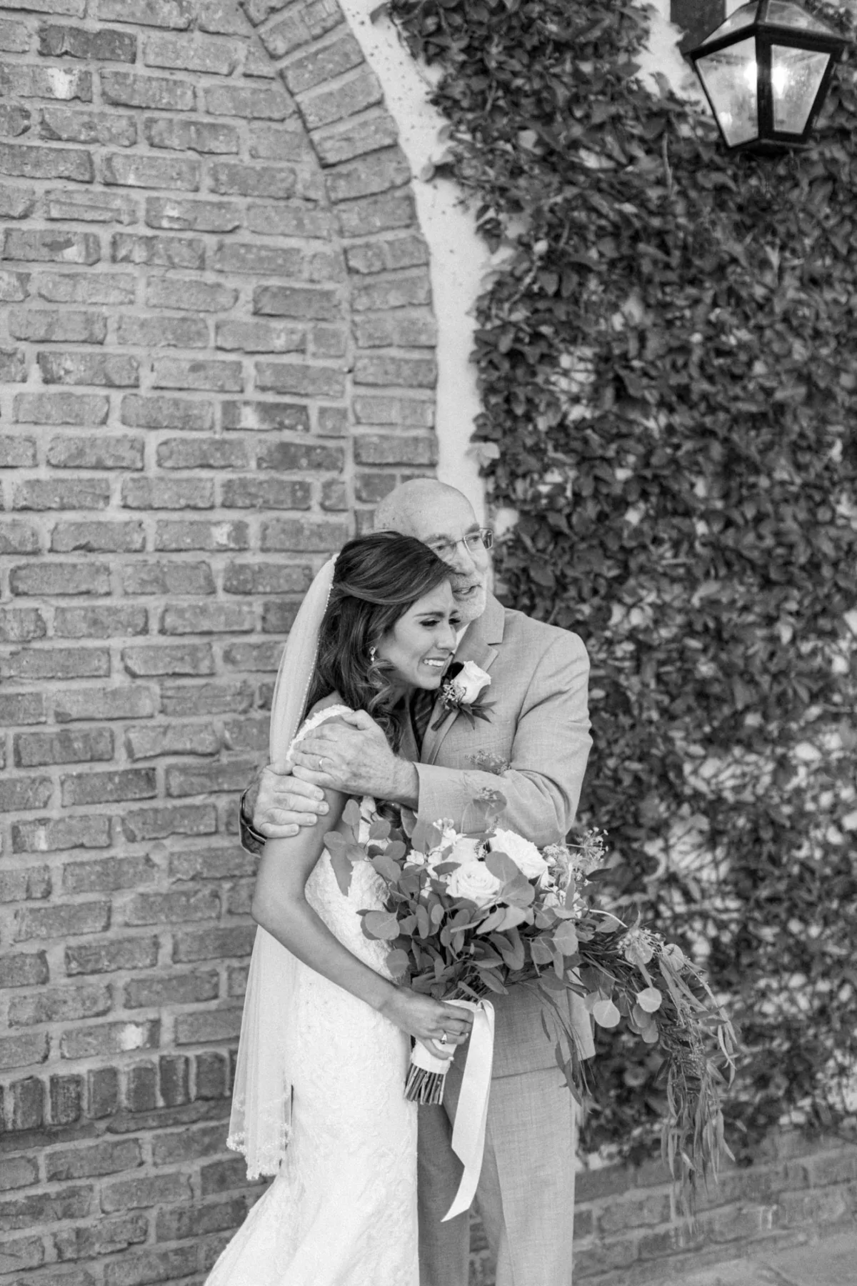 Black and white photo of a bride hugging her father outside a brick wall at Bella Collina, holding her wedding bouquet.