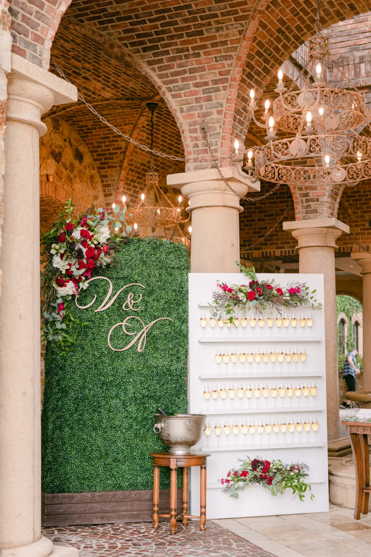 Champagne wall display at Bella Collina wedding venue with greenery backdrop, floral arrangements, and hanging chandeliers.