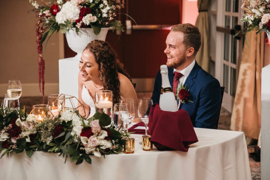 bride and groom sitting at sweetheart table with white, red, and green flowers on display on the table and behind them created by The Flower Studio