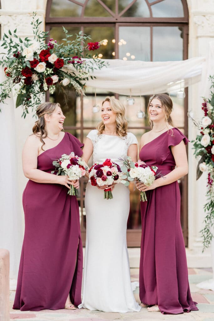 bride and two bridesmaids in plum colored dresses smiling and holding their matching bouquets prepared by The Flower Studio