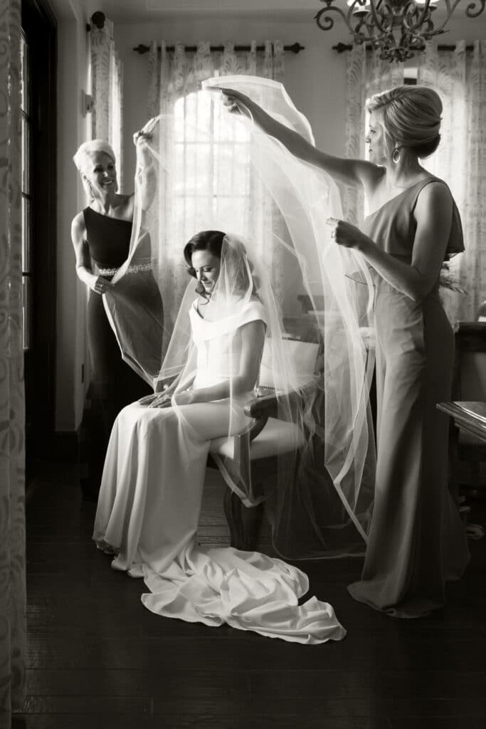 black and white image of bride sitting while bridesmaid and mother bring large veil over her head