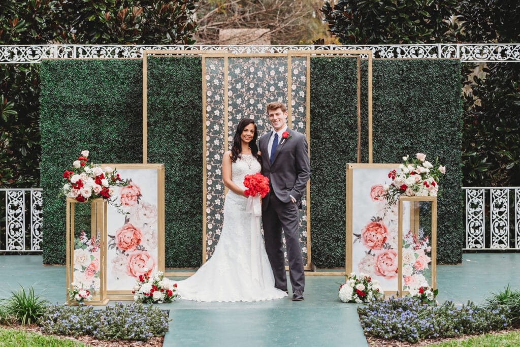 bride and groom standing in front of green vine covered wall with white and red flowers on custom podiums next to them