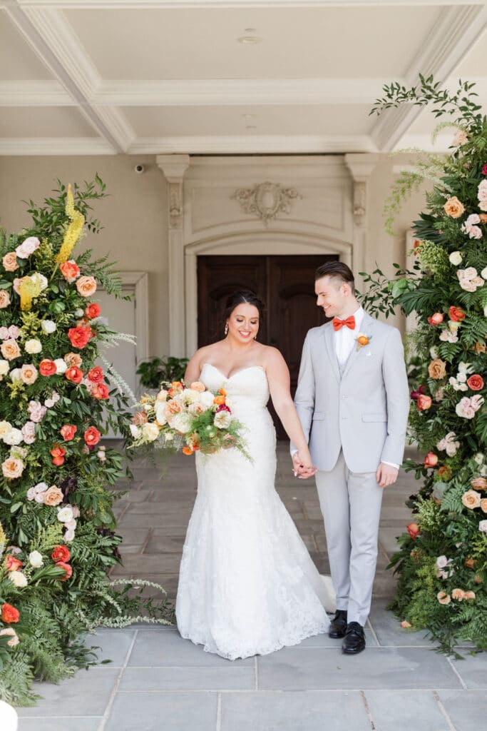 bride and groom holding hands in front of building in between two large floral arrangements