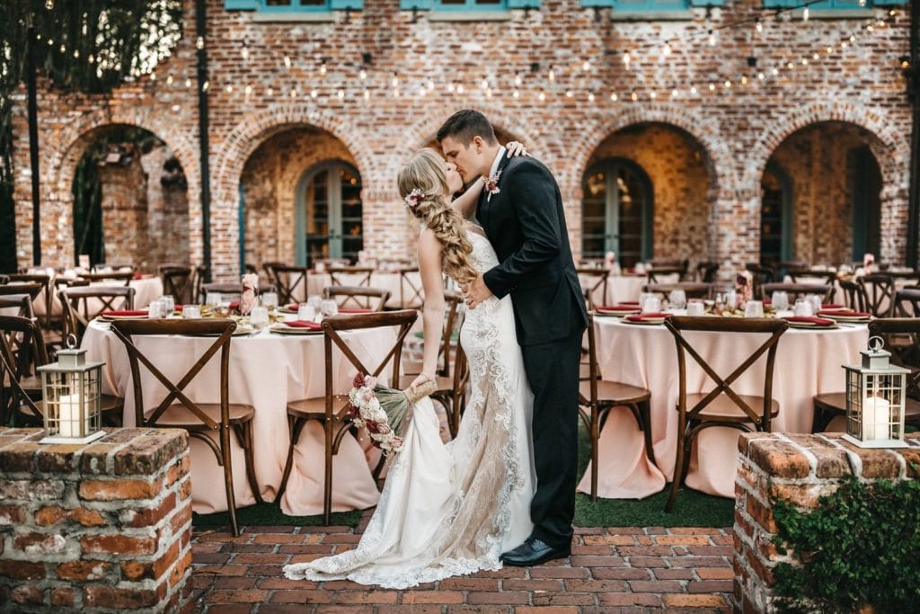 As-You-Wish- Bride and Groom kissing in the middle of table arrangement in front of brick wall