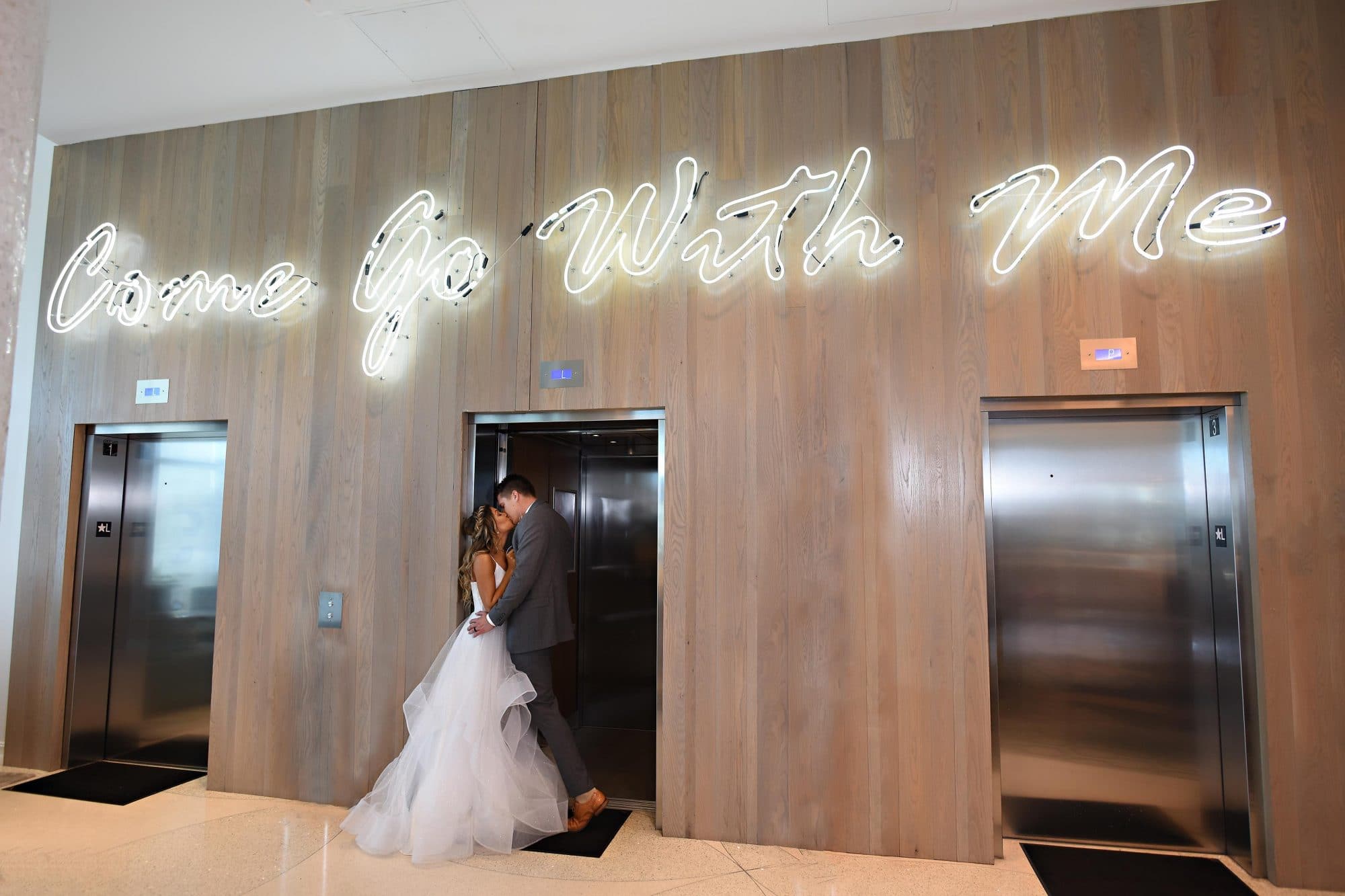 bride and groom kissing in elevator with neon sign above reading "Come Go With Me"
