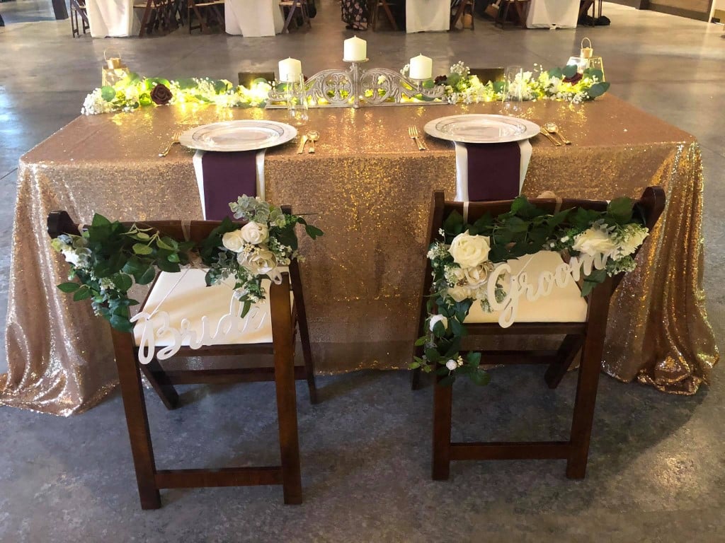 Perfect-Day-Productions-Bride and Groom chairs at Wedding reception with white flowers on a vine and gold sequin table cover.
