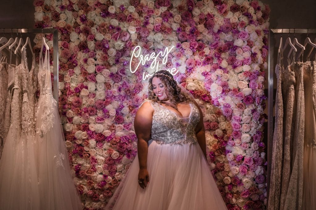 curvy bride in front of pink flower wall with wedding dresses