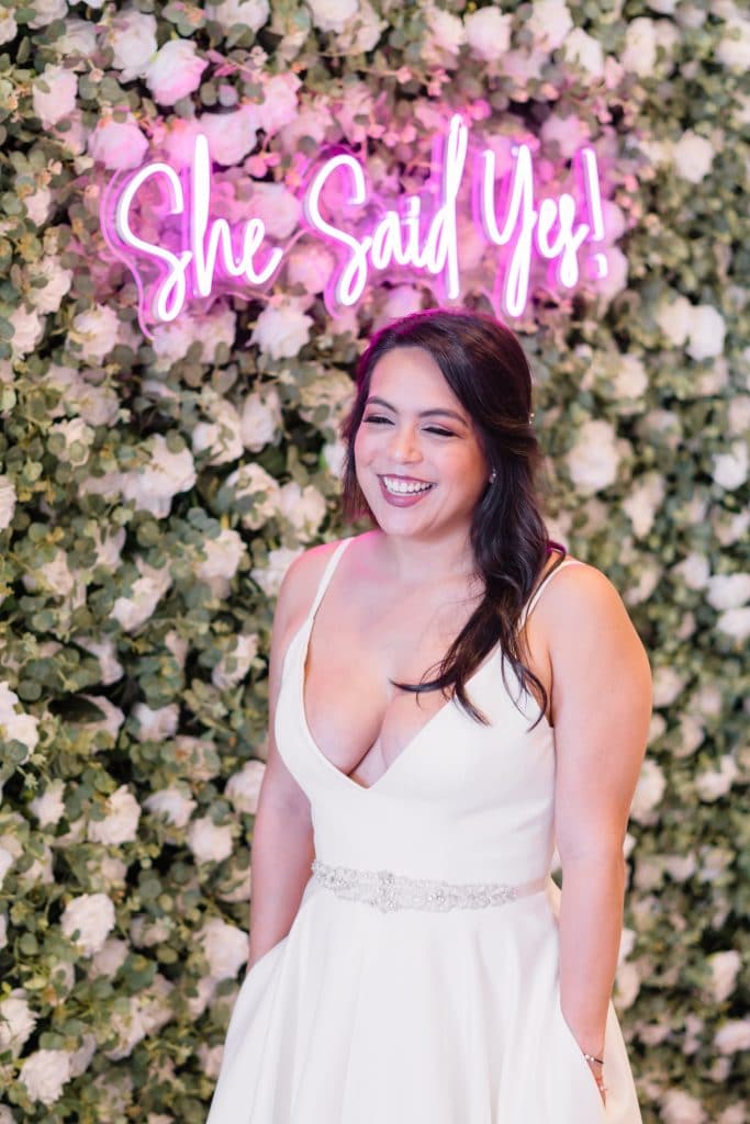 bride with long brown hair in front of white flower wall and she said yes neon pink sign