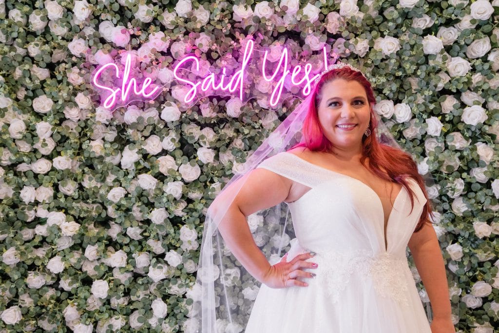 bride with pink hair in front of white flower wall with neon she said yes sign