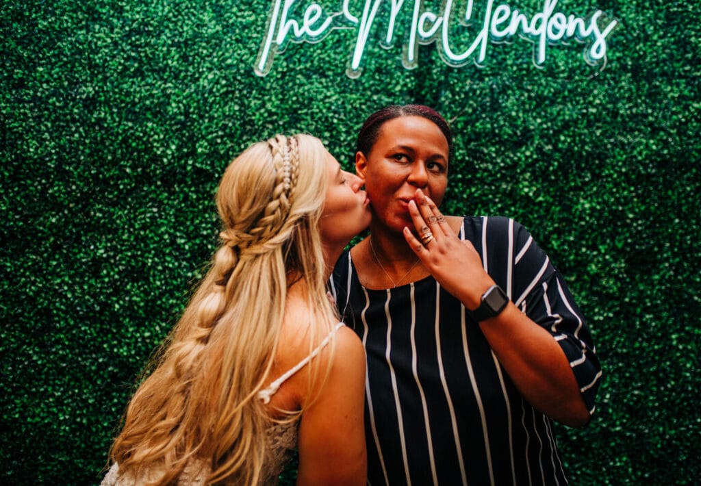 bride with braided blond hair kissing friend on the cheek in front of greenery wall at wedding reception