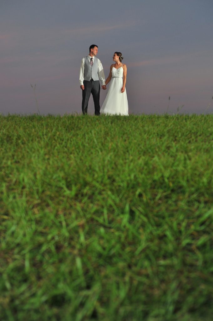 Jennifer Juniper Photography - Bride and groom standing on grassy field at dusk