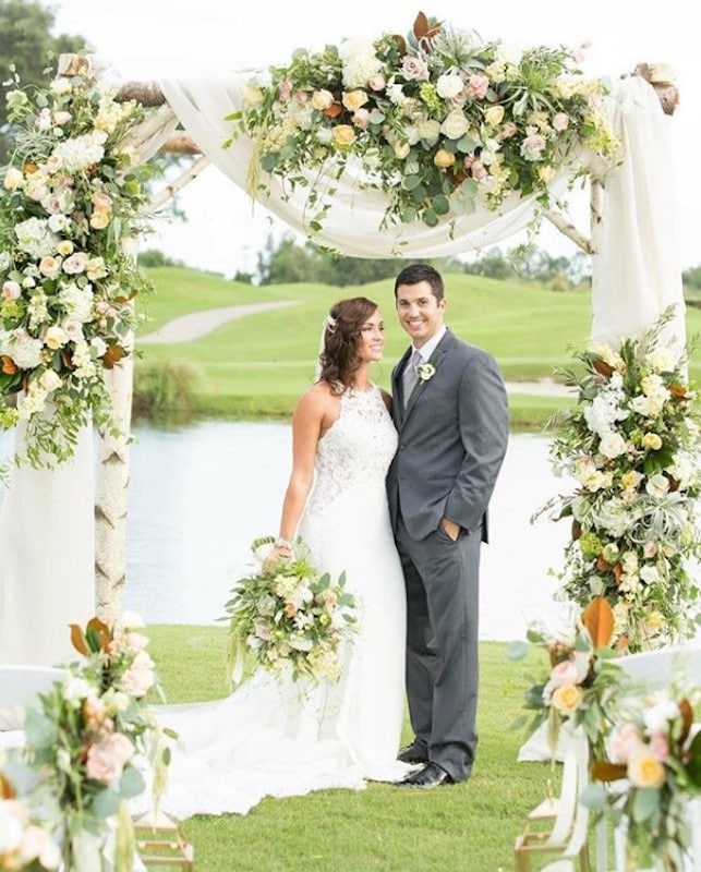 Lowe and Behold bride and groom standing under arch covered in flower arrangements while bride holds matching bouquet