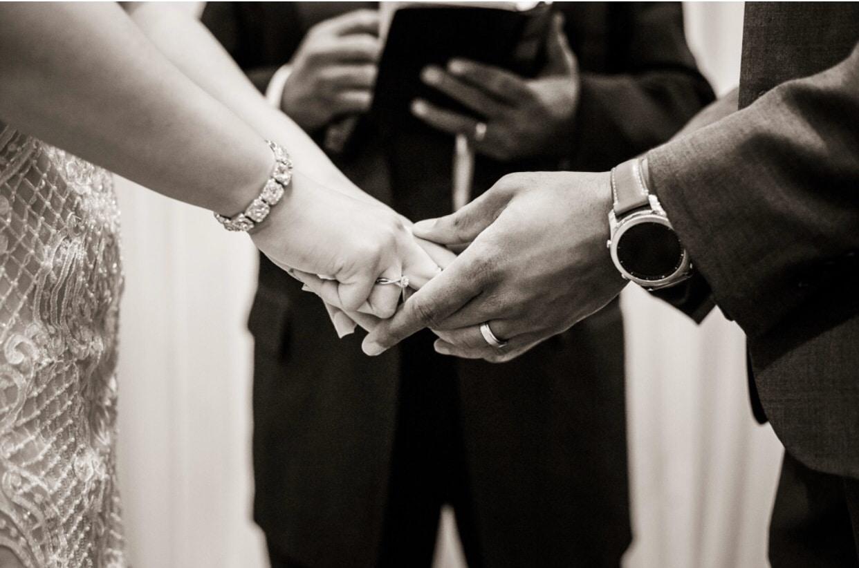 With This Ring, black & white close up of bride & groom holding hands in front of officiant