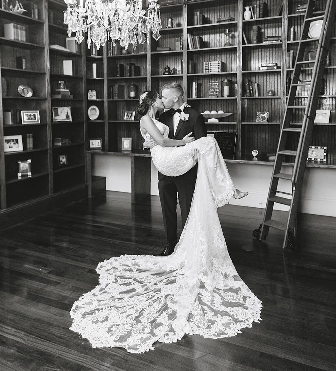Groom holding bride with train on floor kissing in library