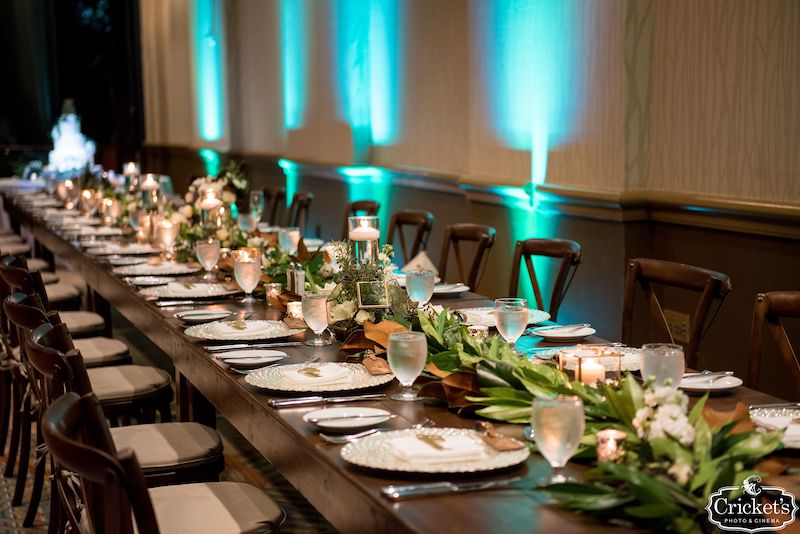 Just Marry long table set up for wedding reception with flower centerpieces and blue uplights along the walls