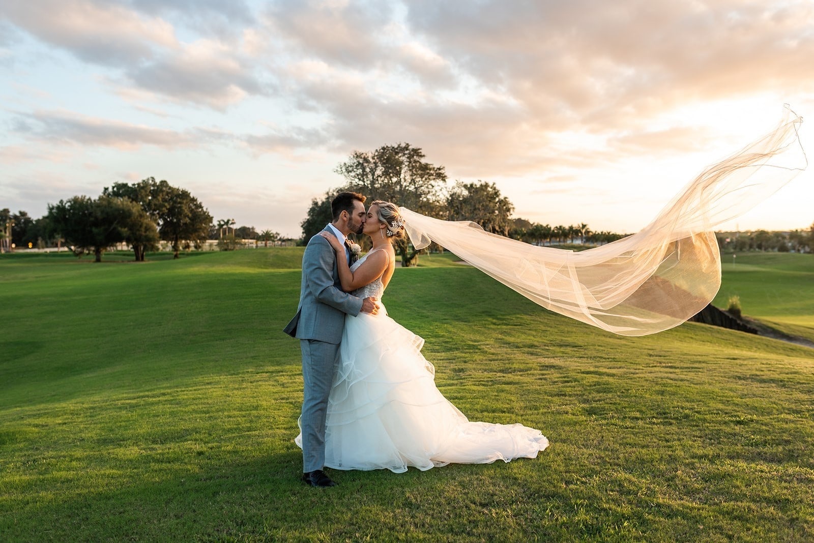 bride and groom standing on golf course during beautiful sunset with brides veil flowing behind her in the wind