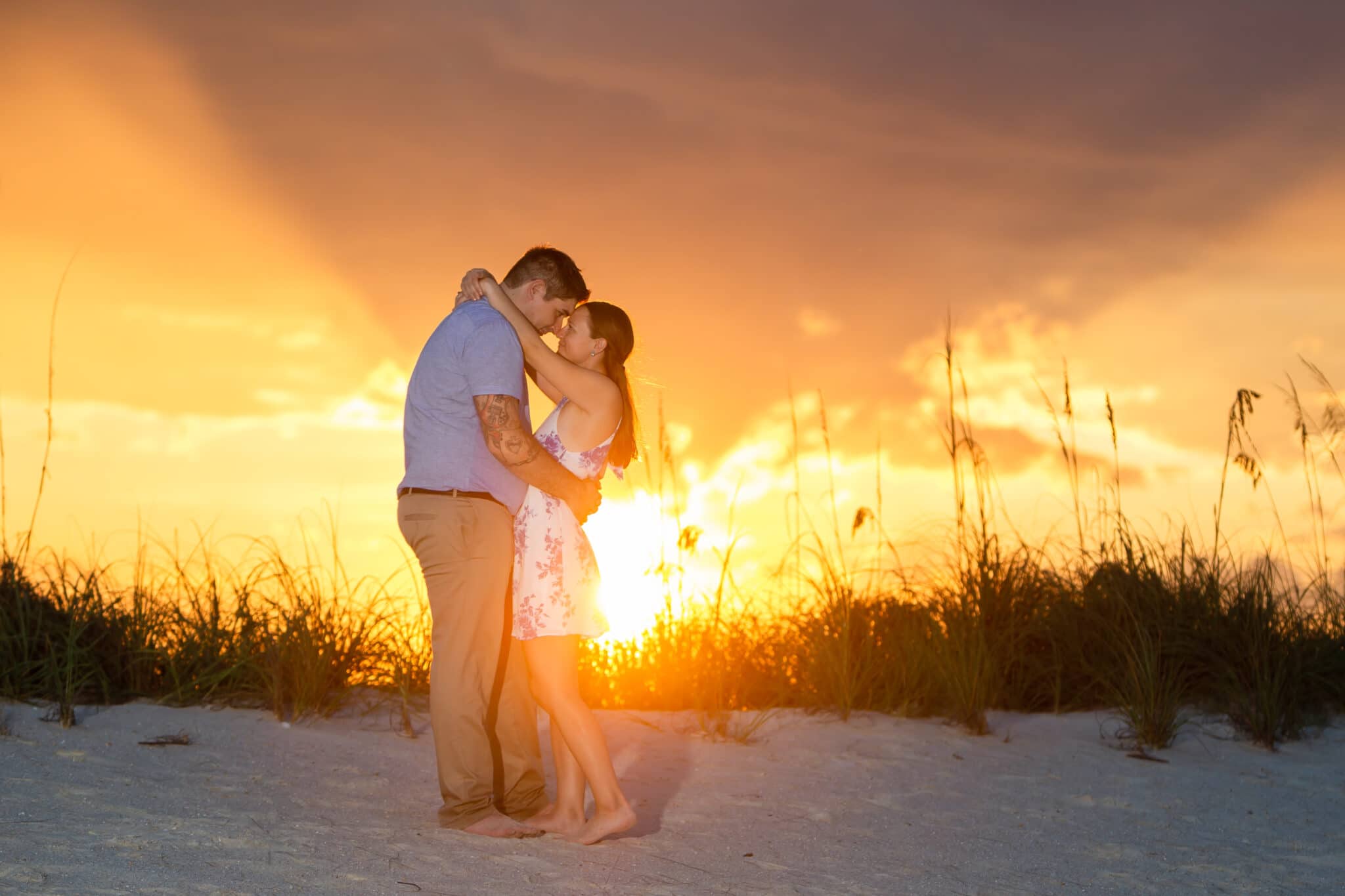 bride and groom standing nose to nose while barefoot on a beach with the dunes and a sunset behind them
