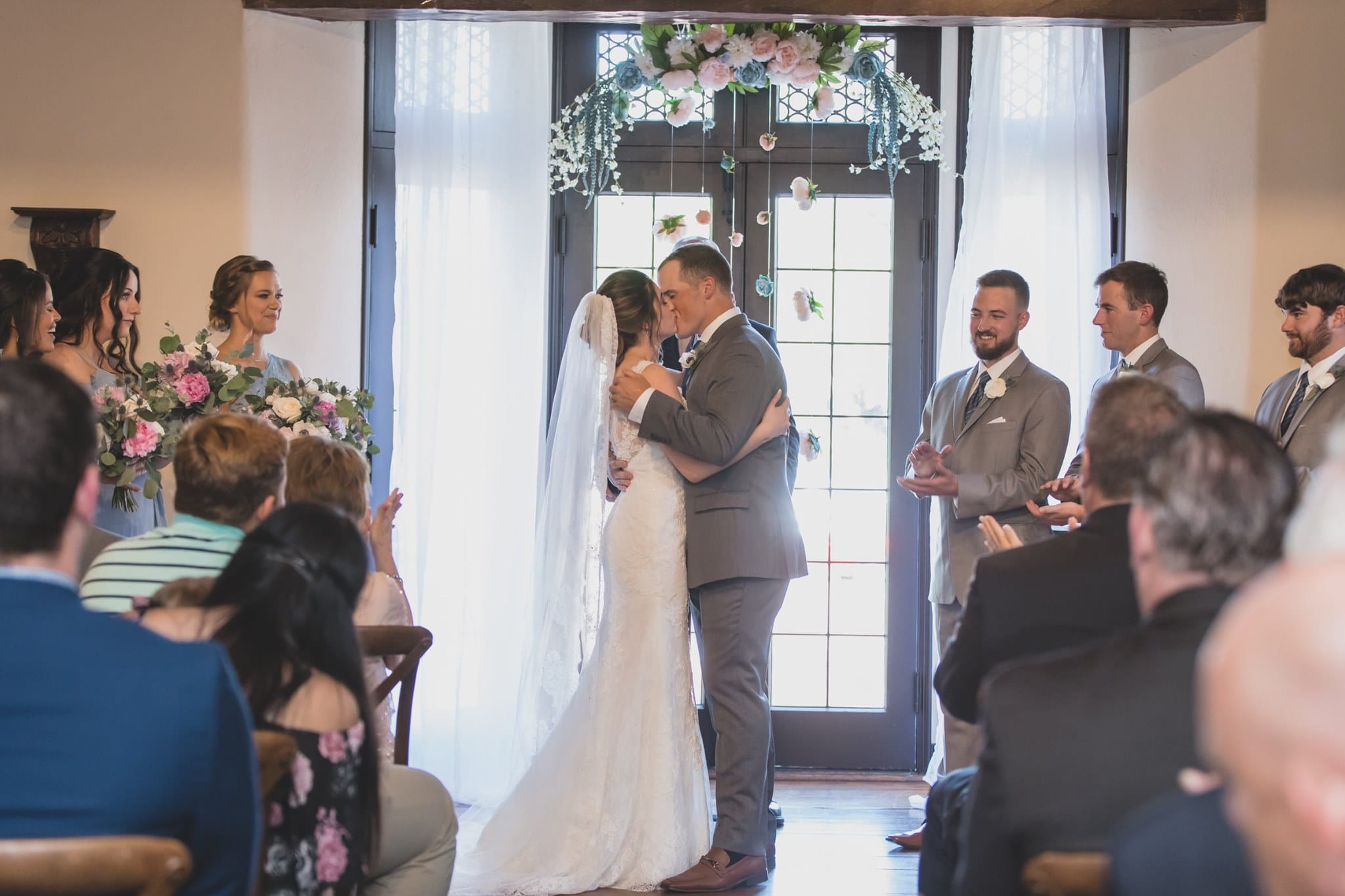 bride and groom kissing during their indoor wedding ceremony while their guests and bridal party cheer them on