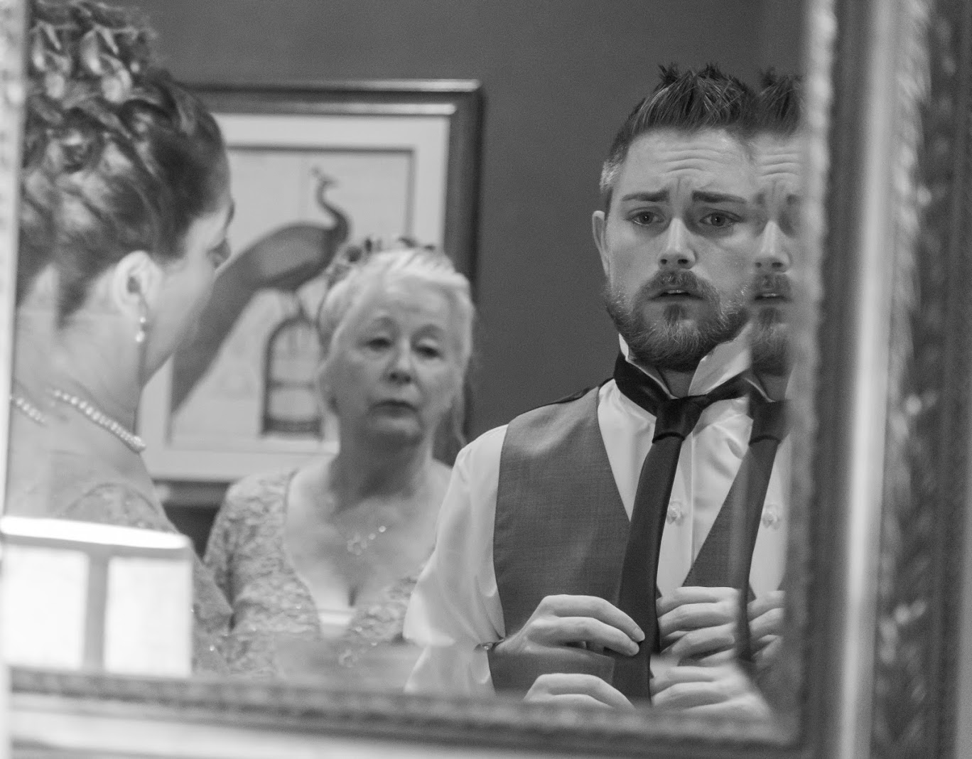 black and white image of groom putting on his tie in mirror with family next to him and behind him