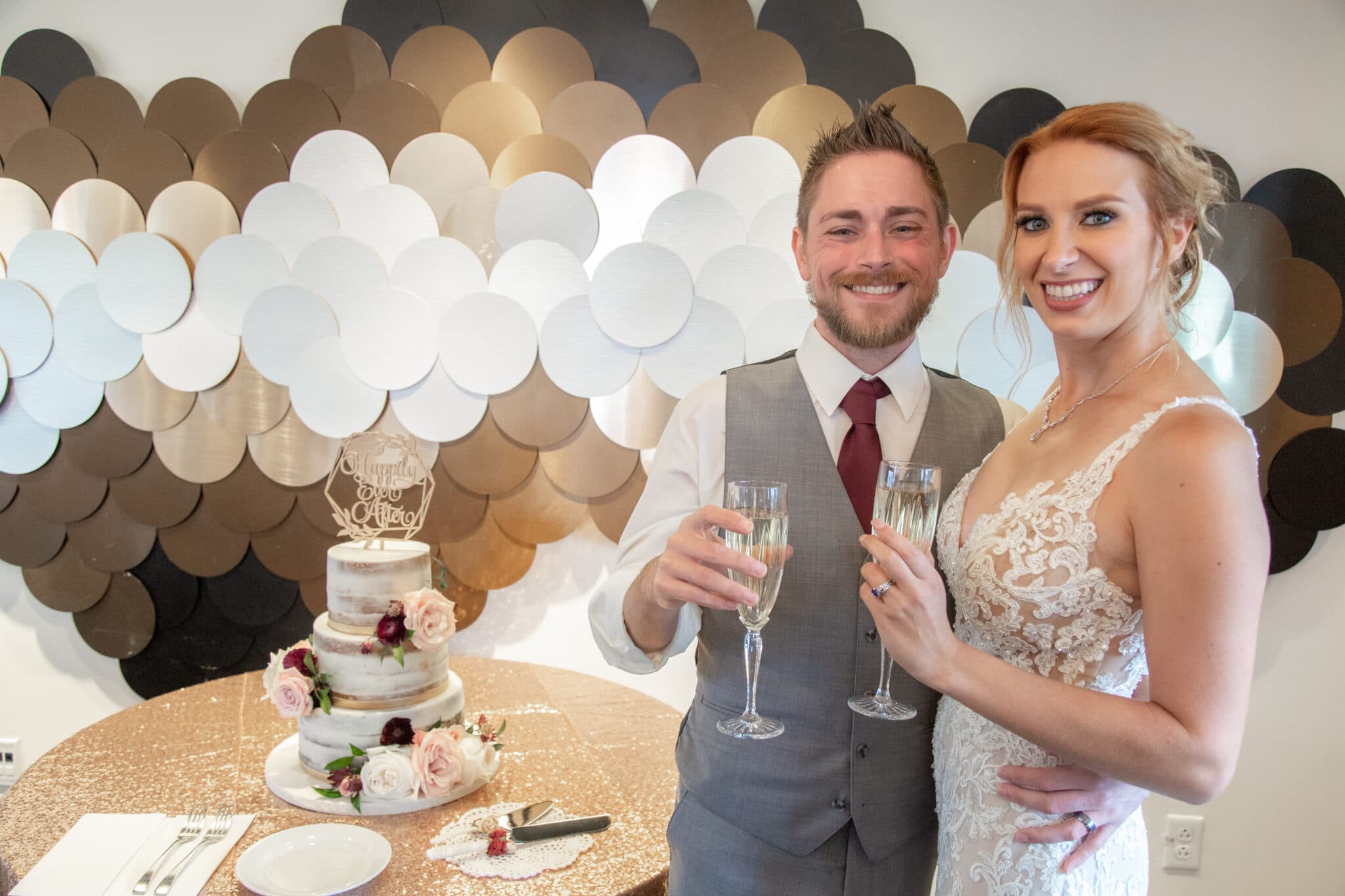 couple holding champagne glasses smiling in front of cake table and gold black and white wall decor