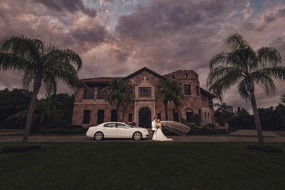 moody and dark shot of bride with floating veil and groom next to classic white car in front of their wedding venue