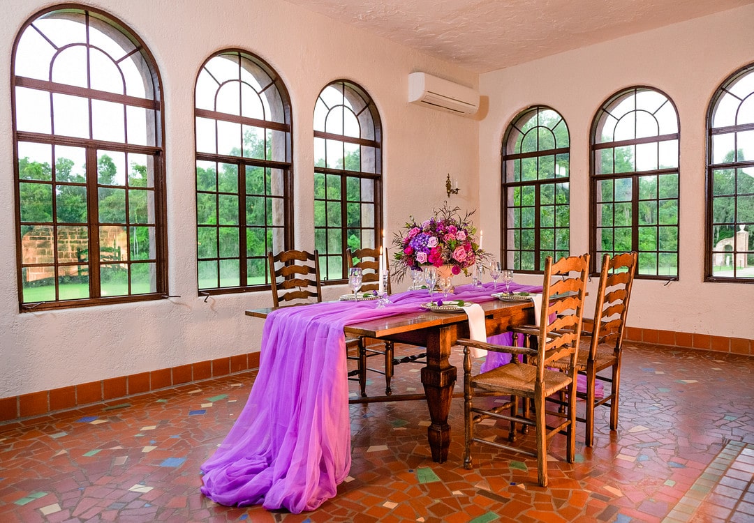 single wooden table in room with fluffy vibrant purple table fabric and big floral arrangement for wedding reception decor