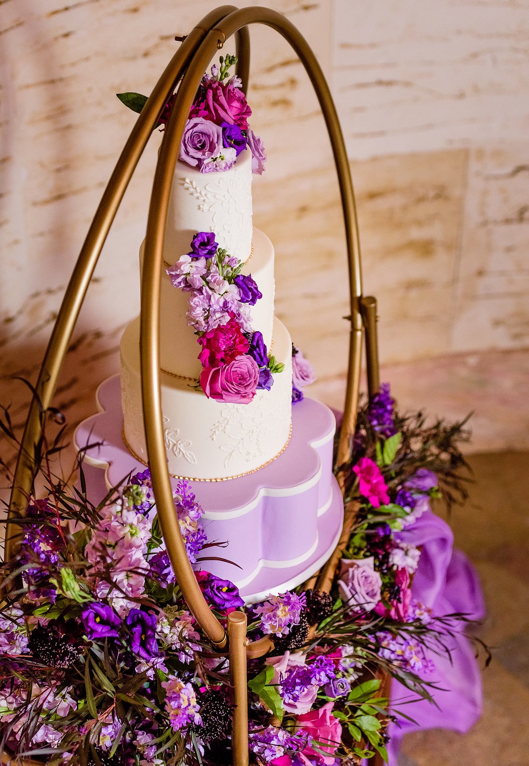 close up of three tiered white wedding cake decorated in matching flowers on golden hoop cake stand surrounded by matching flowers