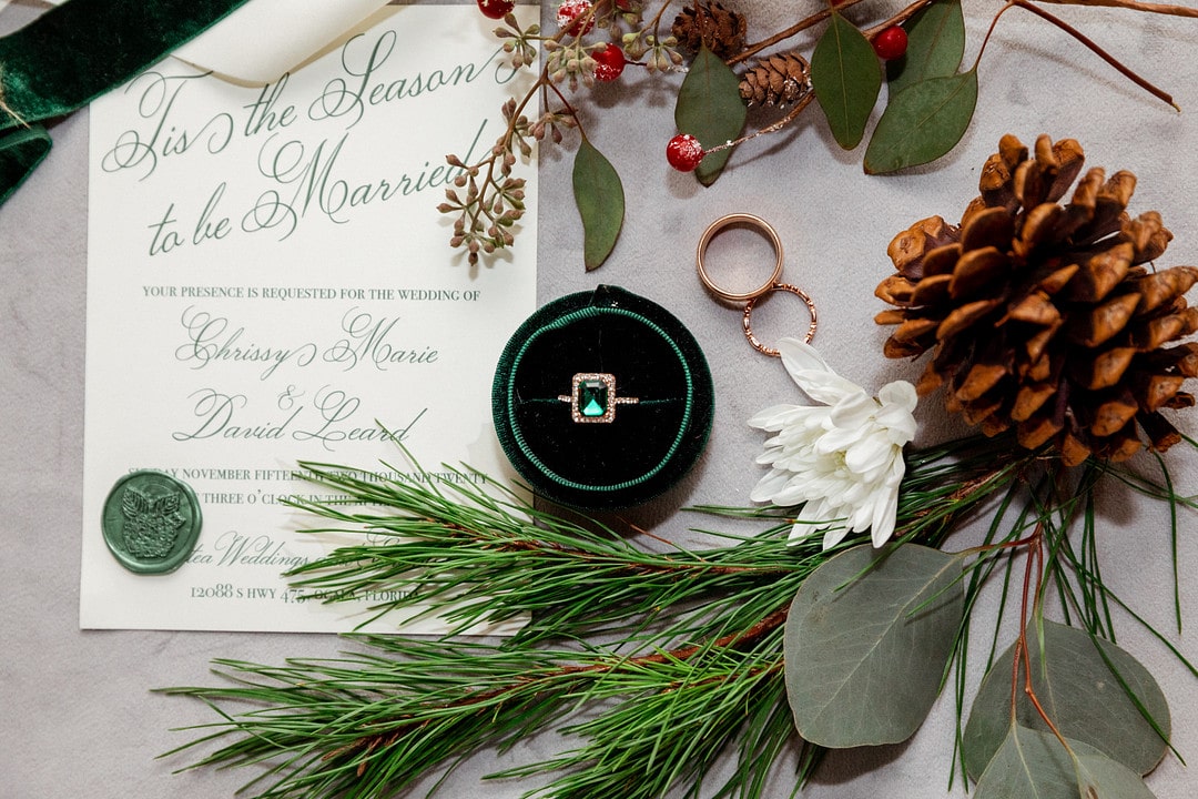 pine cone and sprigs of pine tree around velvet green ring box with square emerald green wedding ring next to wedding invitation