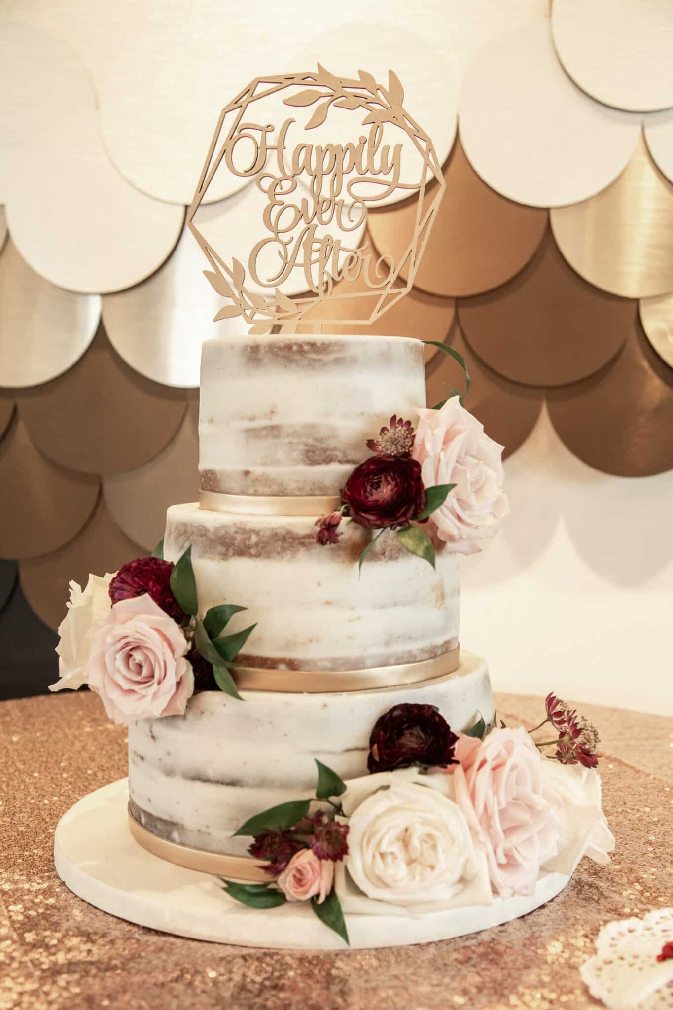 naked frosted three tier wedding cake with gold ribbon and cream pink and maroon flowers with a custom cake topper