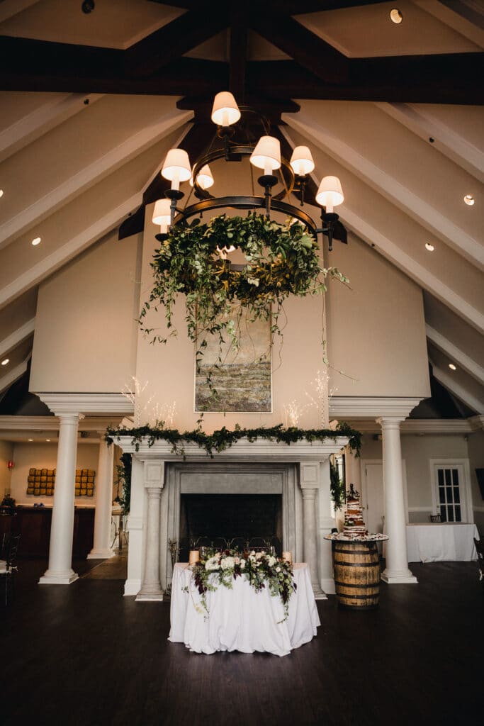 sweetheart table set in front of a large fireplace, with mantle and chandelier decorated and the wedding cake on a barrel behind the table
