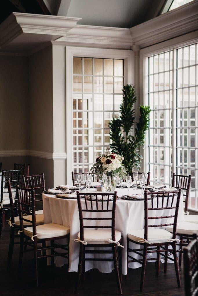 table setup for wedding reception in front of large windows, with flower centerpieces and tall potted plant in background