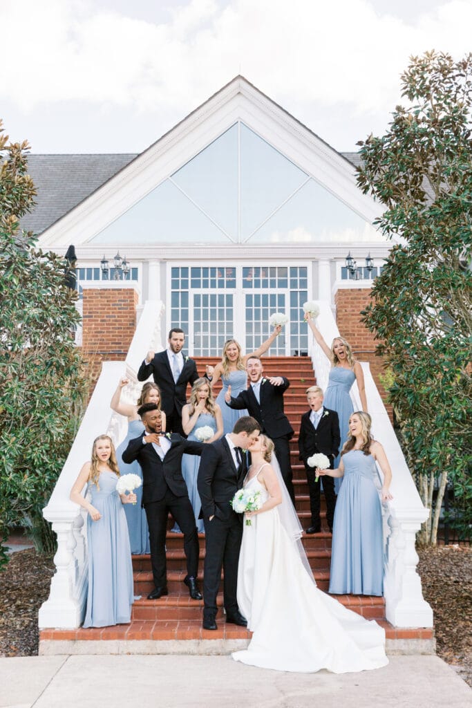 bride and groom standing on bottom of stairs kissing while bridal party celebrates