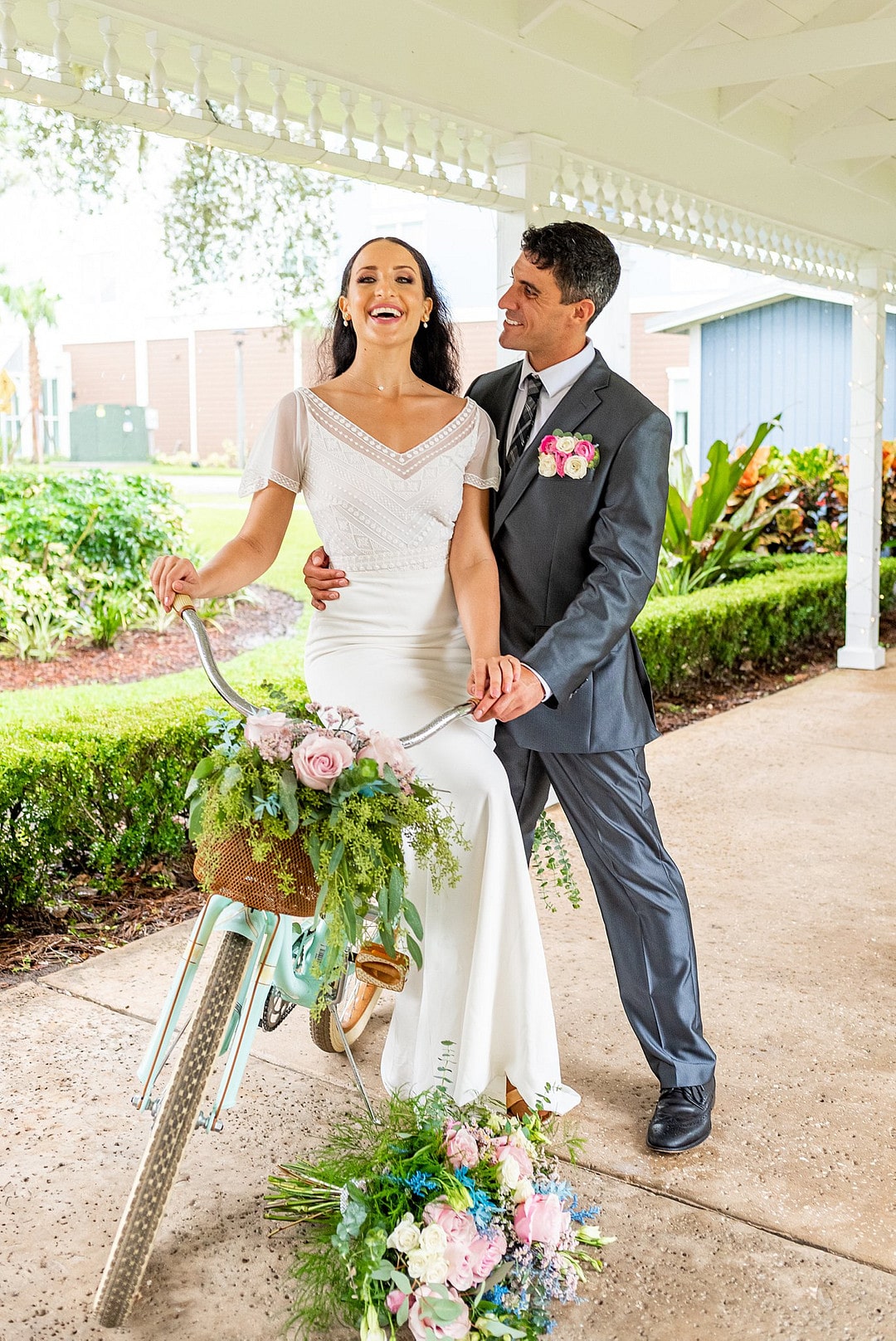 bride sits on vintage bicycle decorated with flowers as her bouquet sits on the ground in front and groom stands behind her smiling at her
