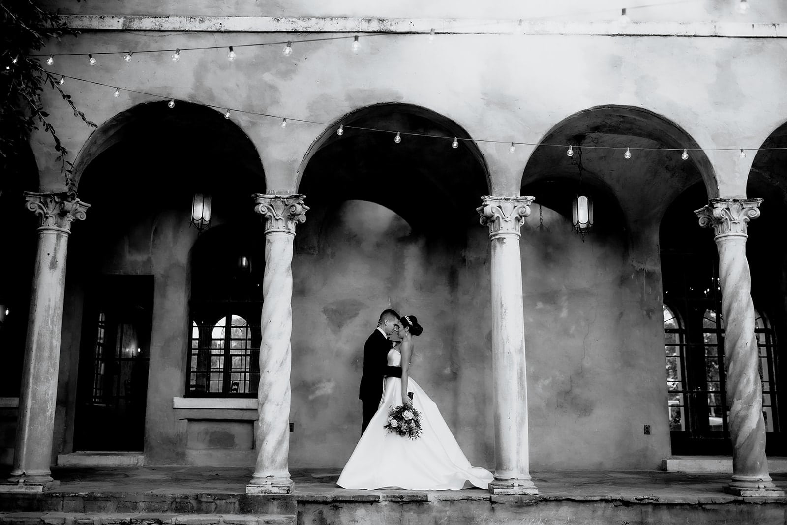 bride and groom stand touching foreheads outside under covered patio between two columns with market lights hanging at the top of the picture outside
