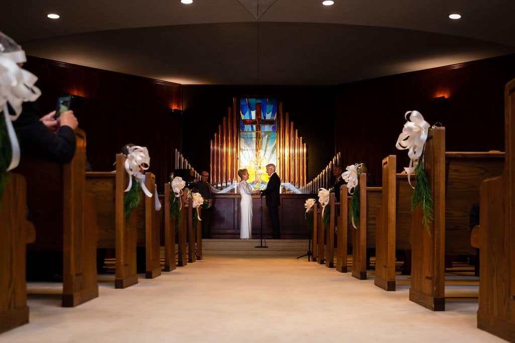 long shot of bride and groom at wedding ceremony about to be married in the front of a chapel and decorated pews with white bows at the ends