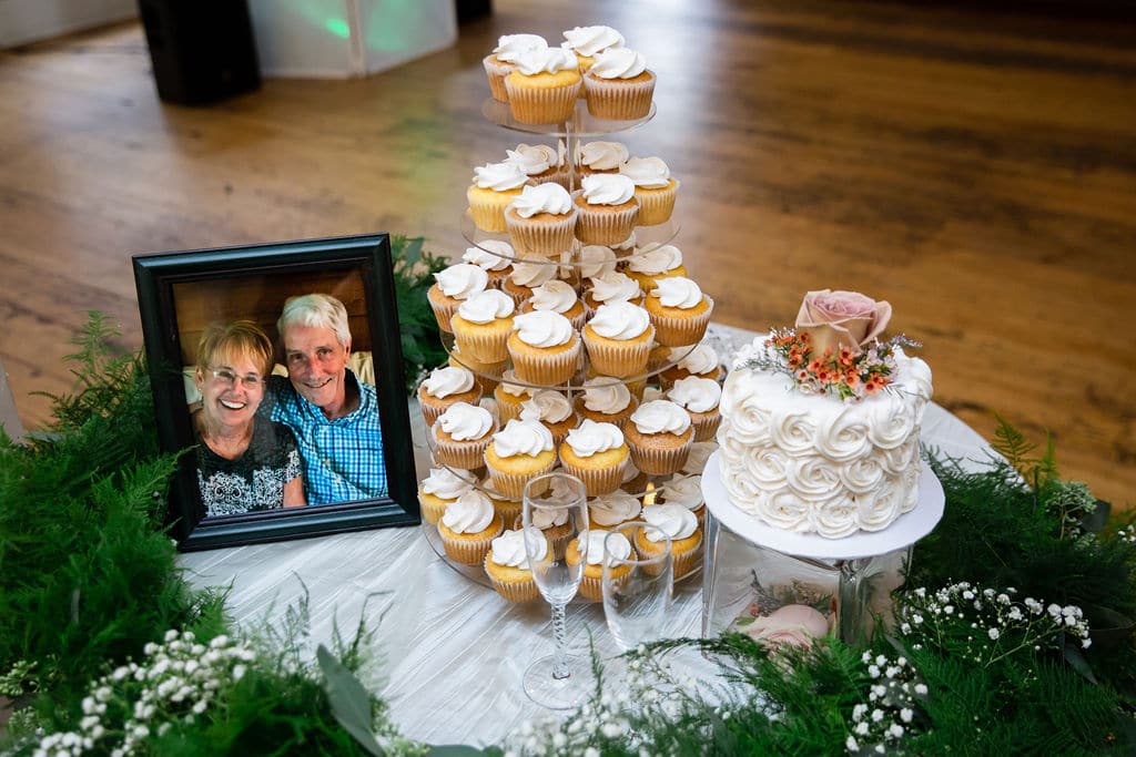 wedding cake table with one tier buttercream flower wedding cake with cupcake tower next to it with picture of couple on table and greenery