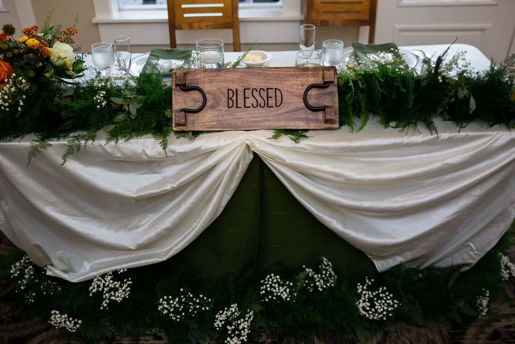 picture of sweetheart table at wedding reception with long run of green garland on top of table and a wooden blessed sign in front in the middle with arrangements of babys breath in front of the table on the floor