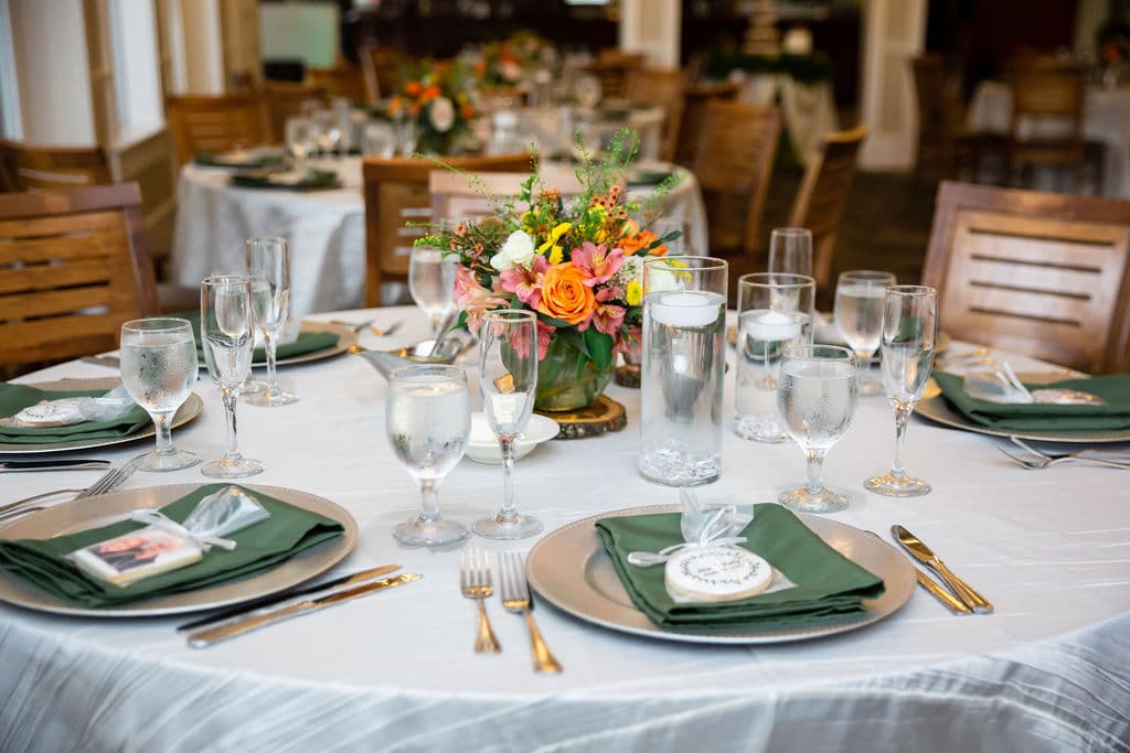 close up shot of wedding reception table with tall vases filled with water and floating candles a small floral arrangement in the middle and chargers at each seat set with green napkins and individually wrapped cookies on top