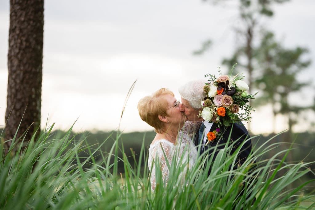 bride holds bouquet around grooms neck and they kiss behind tall grass
