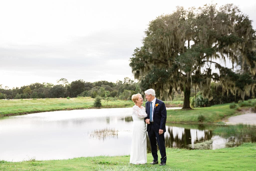 married couple stands in front of body of water on golf course and a big tee smiling at each other for wedding day pictures